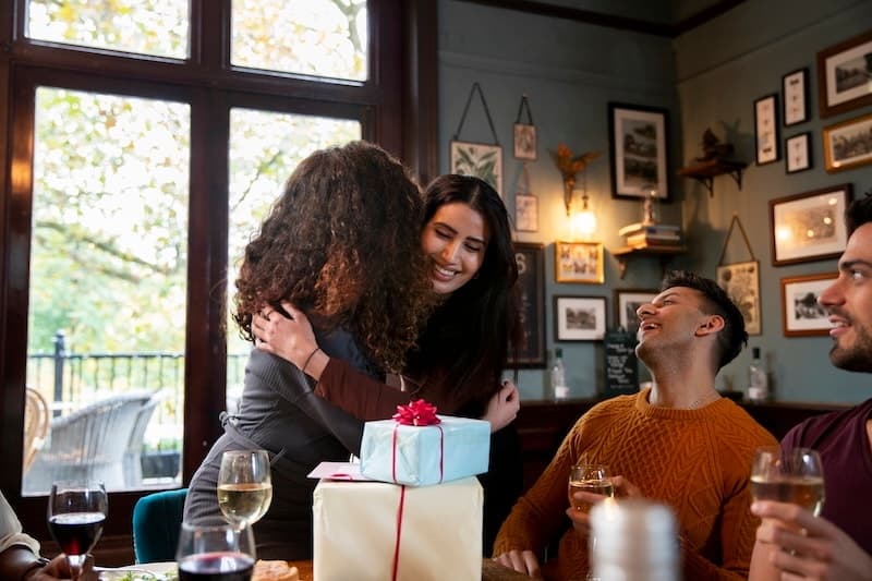 Two friends embracing over a wrapped gift at a restaurant table