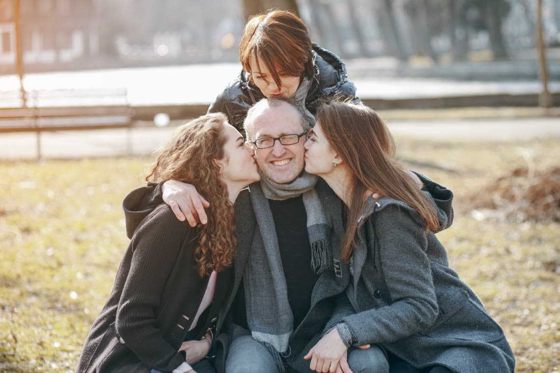 A father in a park being kissed on both cheeks by his grown daughters