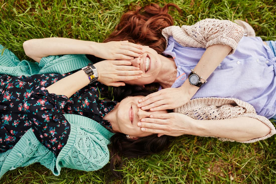 Two friends lying head to head on the grass, covering each other's eyes