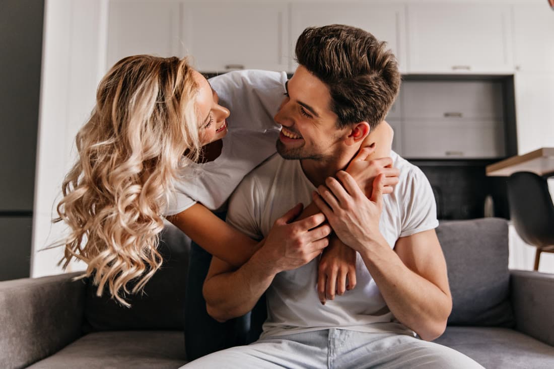 A wife leaning over the back of the sofa with her arms around her husband