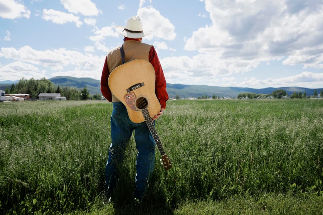 A man standing in a field with an acoustic guitar on his back